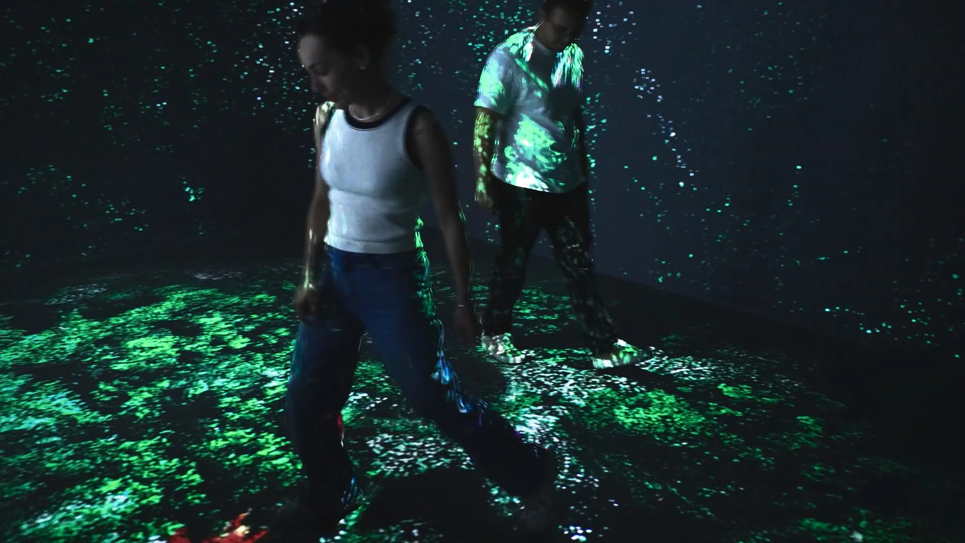 Two visitors on a floor of dense green particles, more particles rising on the walls