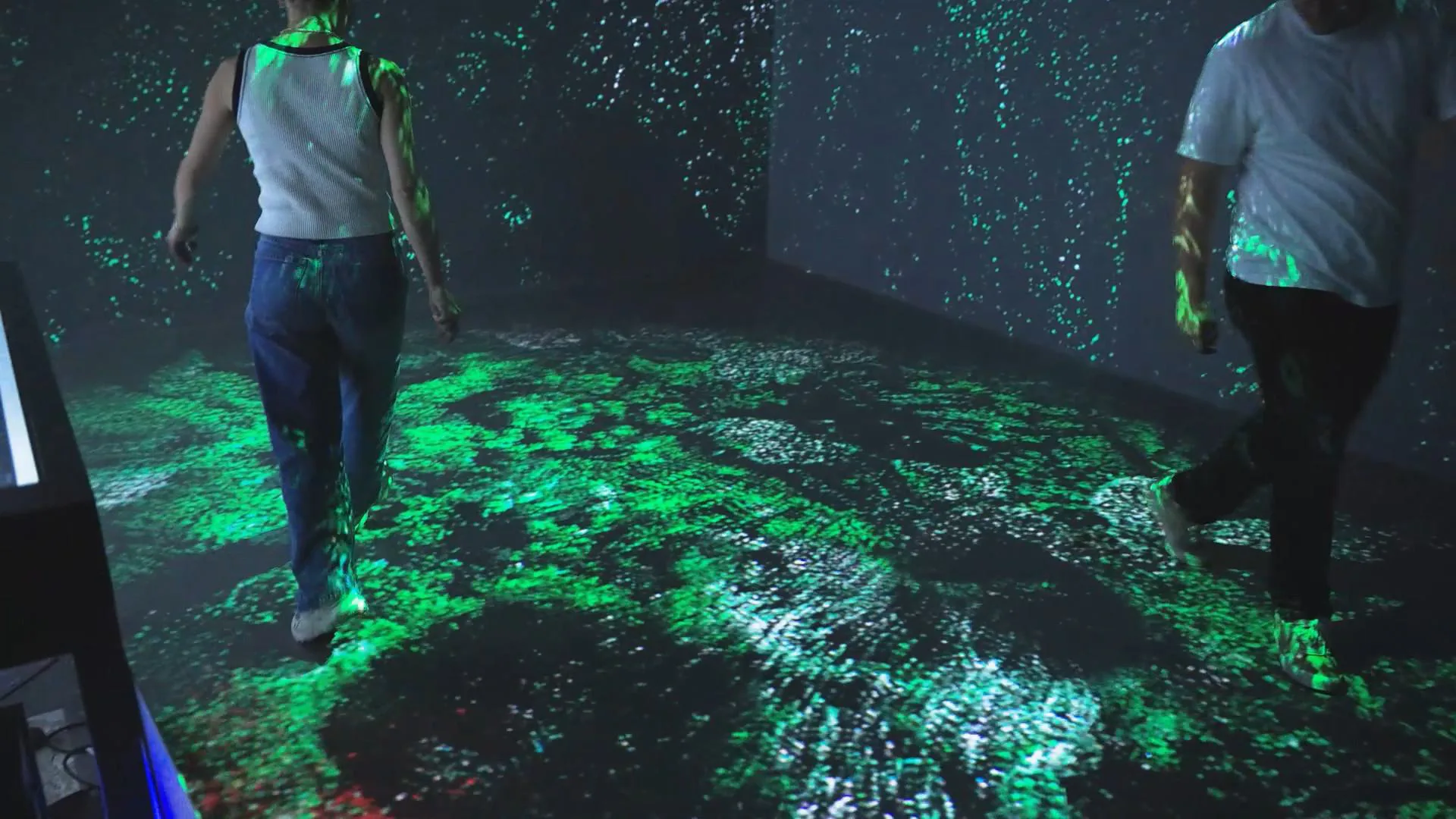 Two visitors walking across a floor of green particles that scatter around their steps, the same particles drifting up the walls