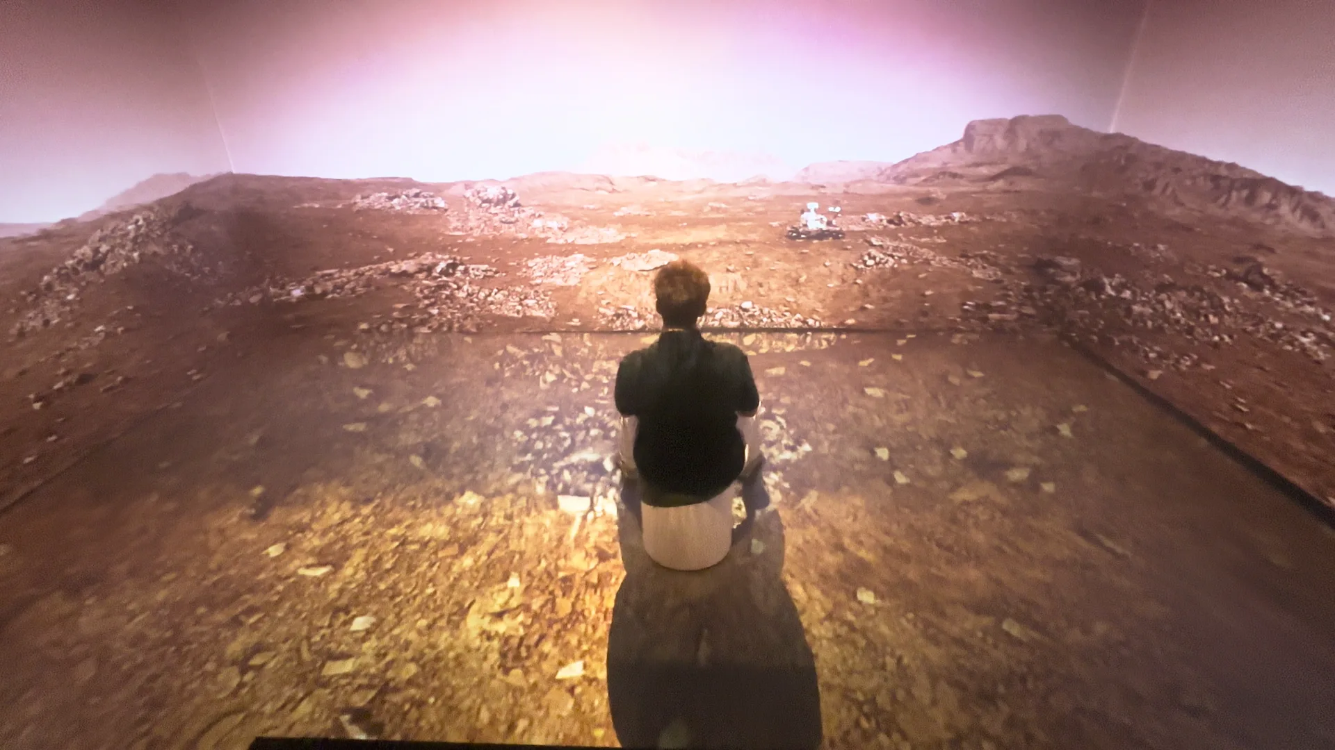A visitor seated alone inside the Martian surface, the rover visible on the far wall