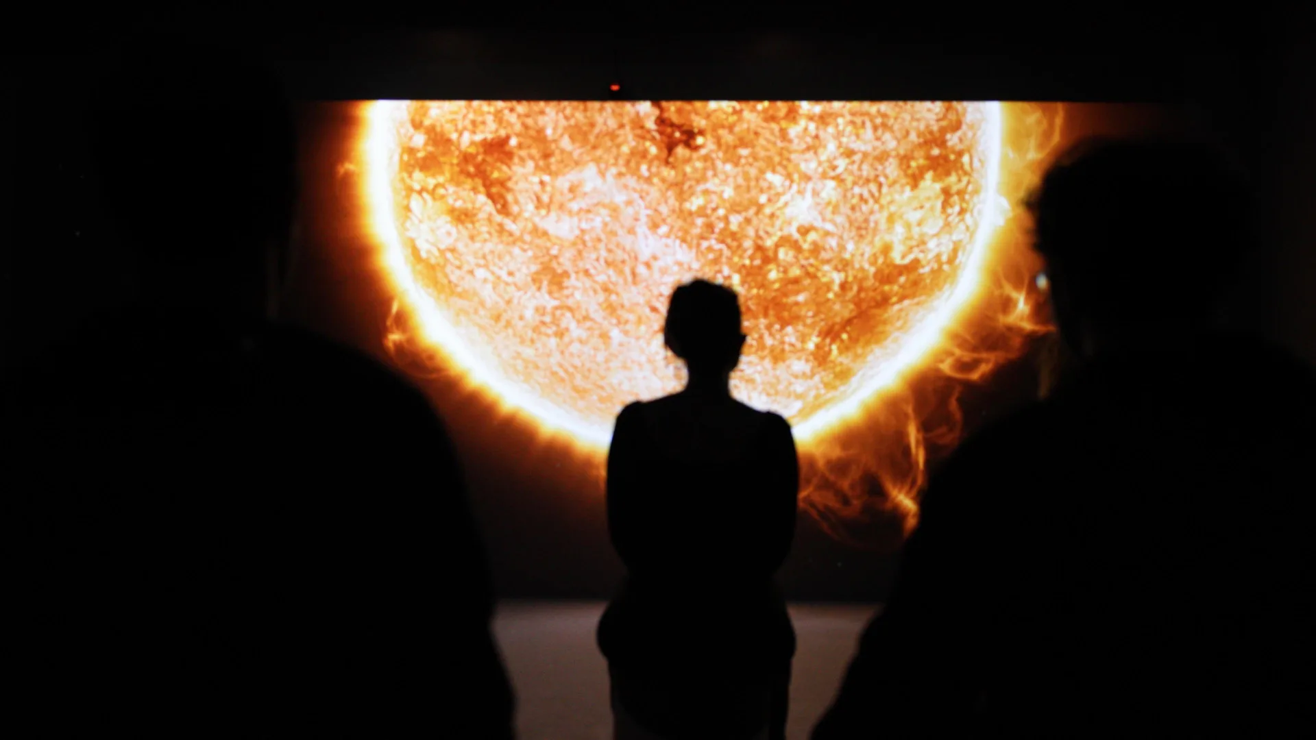 Three visitors in silhouette in front of the Sun, projected wall-to-wall across the immersive room