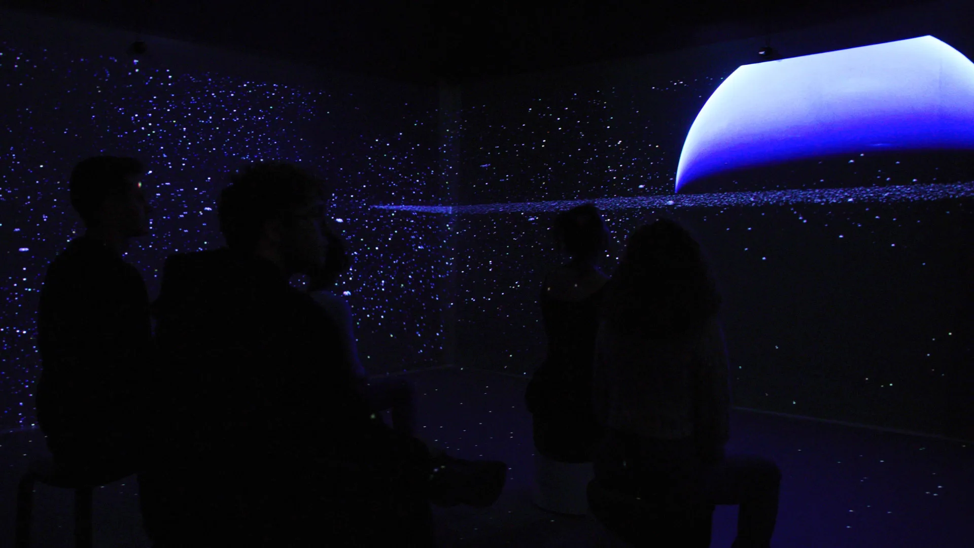 An audience seated in a dark room as Neptune's curve and a field of ice fragments wrap the walls